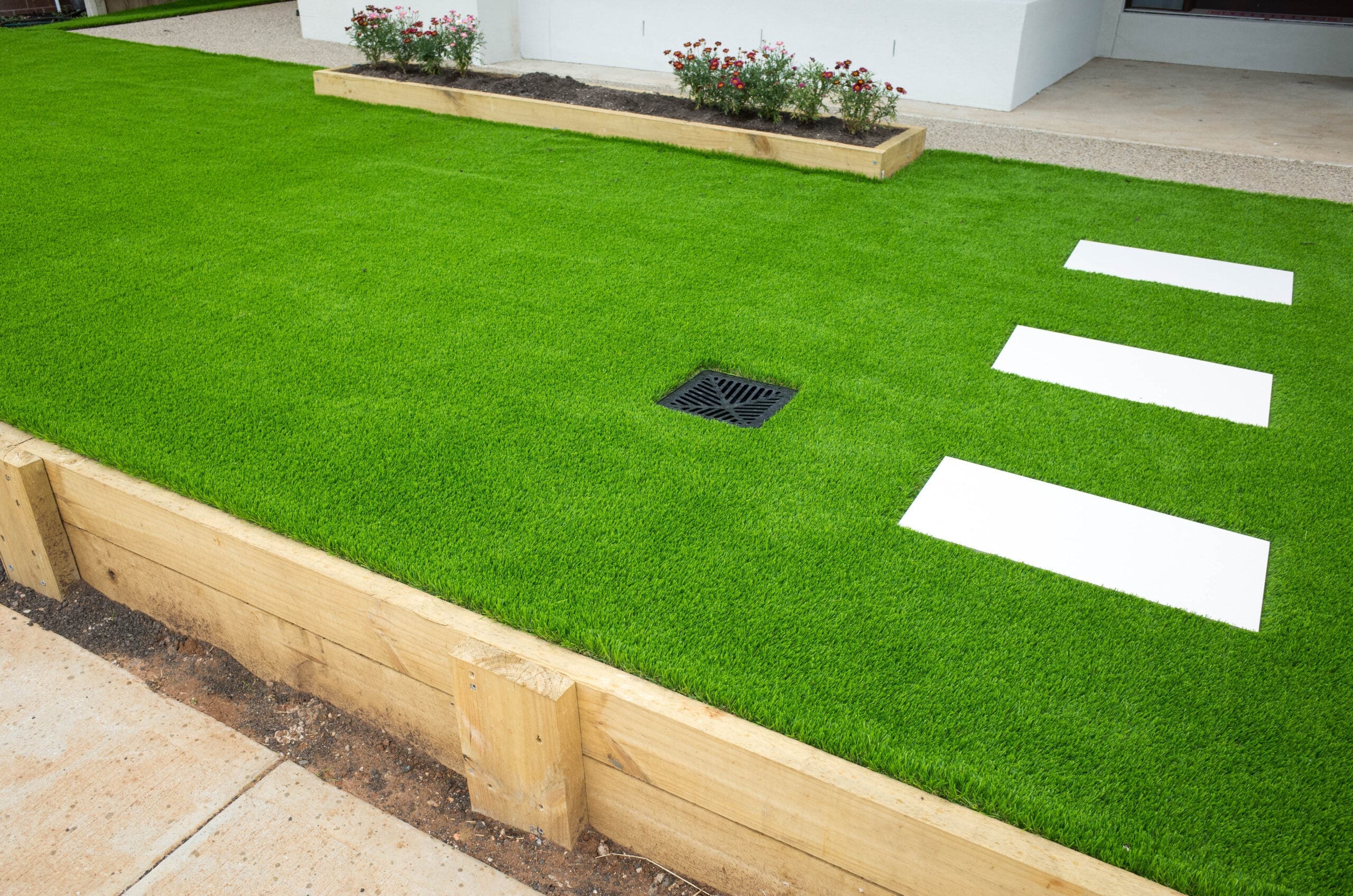 Artificial turf lawn with white stepping stones, wooden retaining borders, and a drainage grate.