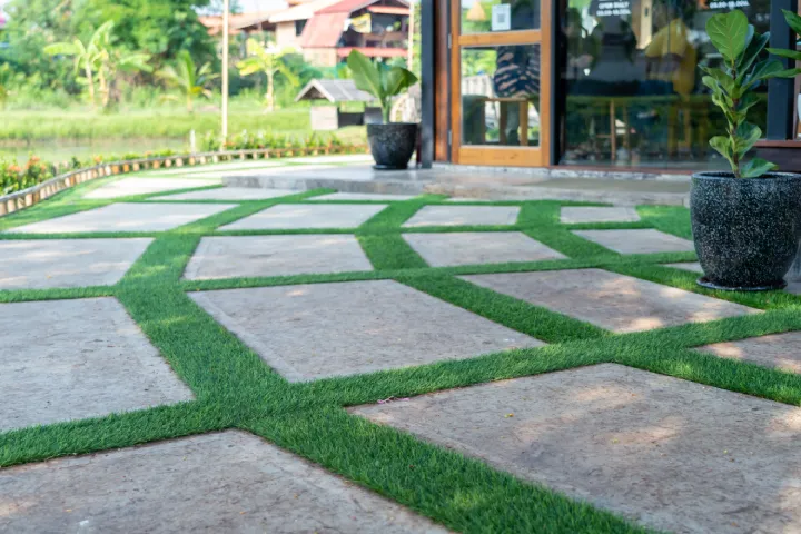 Large concrete pavers separated by strips of artificial green grass near a building entrance.